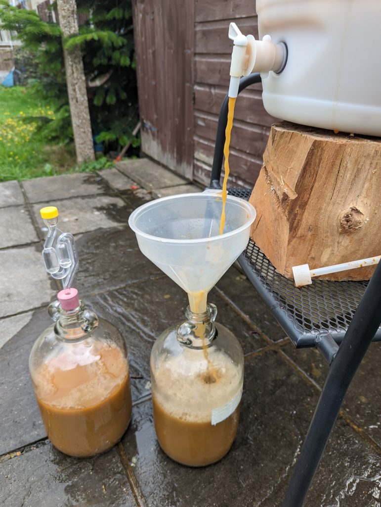 Two glass demijohns, one with a funnel in, being filled with murky brown liquid from a white butt.